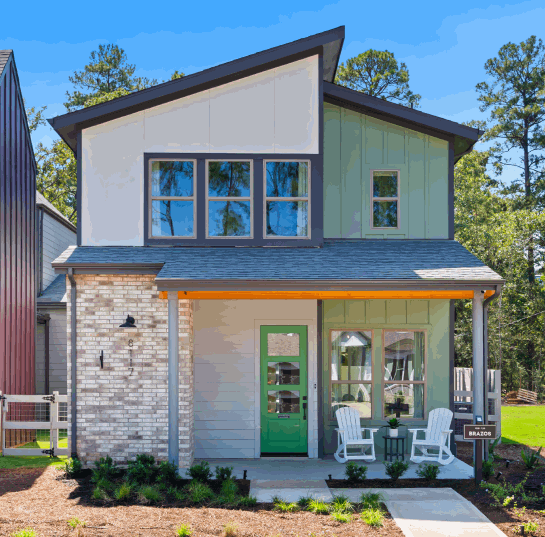 Modern two-story house with a green front door, white chairs on the porch, and large windows. This gem showcases brick and siding, surrounded by trees—perfect for those exploring new homes in Montgomery County, TX near The Grazery restaurant at Two Step Farm.
