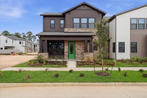 Two-story modern house with brown siding, brick accents, a green front door, and a covered porch, situated on a freshly landscaped lawn in a suburban neighborhood.
