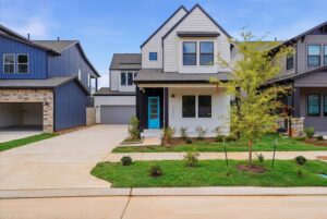 Modern two-story house with a front porch, blue door, small landscaped yard, and driveway, located between two other houses in a suburban neighborhood.