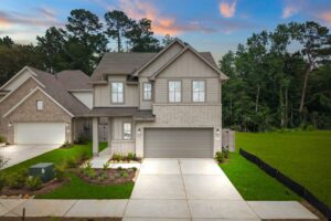 Two-story suburban house with gray brick exterior, attached double garage, manicured lawn, and a wooded area in the background under a colorful sunset sky.