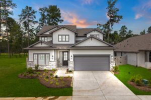 Two-story modern suburban house with gray siding, brick accents, double garage, and manicured yard, set against a sunset sky and surrounded by trees.