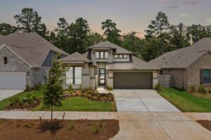 A modern single-story house with a two-car garage, landscaped front yard, and concrete driveway, situated between similar homes with trees in the background at sunset.